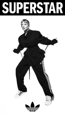 Black-and-white fashion shot of a woman in a dark jacket and Adidas track pants posing against a white backdrop with the Adidas logo.