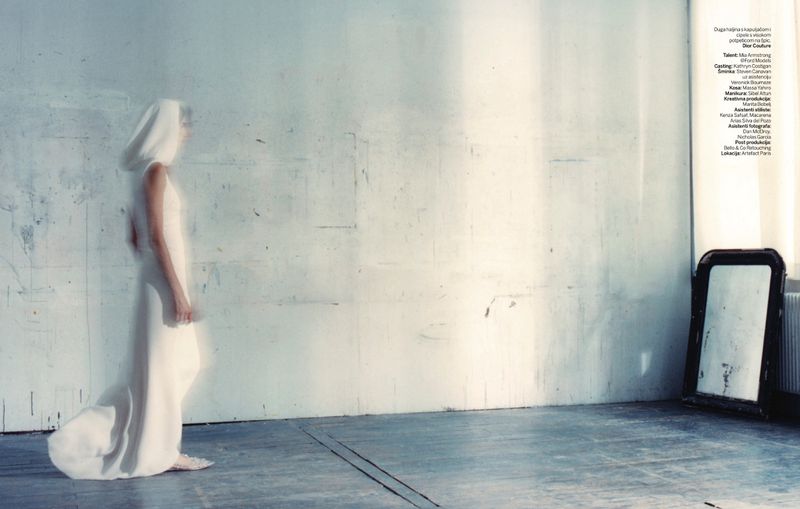 Woman in a white hooded gown walks across a gray floor, long exposure, creating a blurred effect, with a distressed mirror in the background.