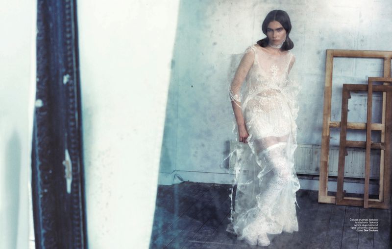 A slender woman poses in a flowing white dress in a photo studio. The composition includes an old wooden frame in the foreground.