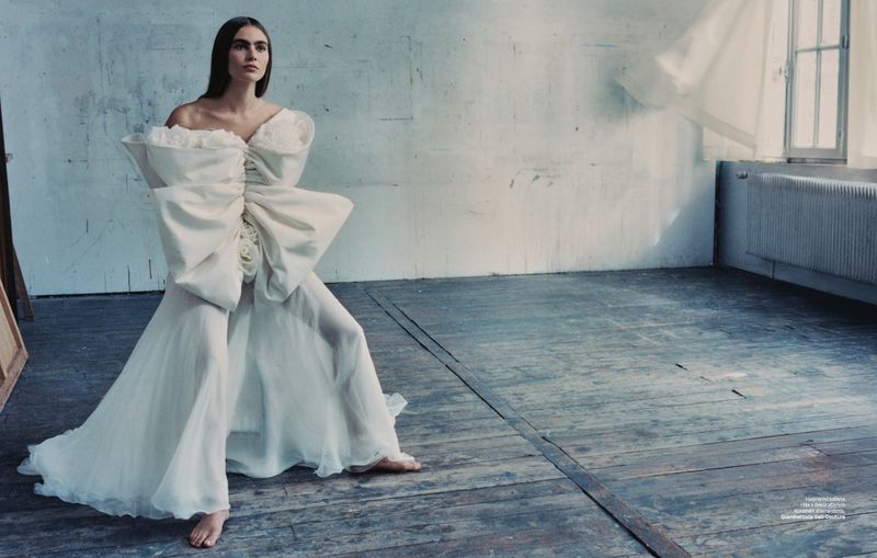 A woman in an oversized white gown poses barefoot in a sunlit room with weathered floors, showcasing fashion and editorial style.
