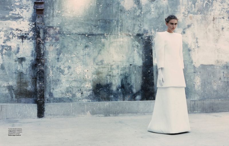 Fashion editorial shot of a model in a white dress and gloves posing against a weathered grey and white wall backdrop.