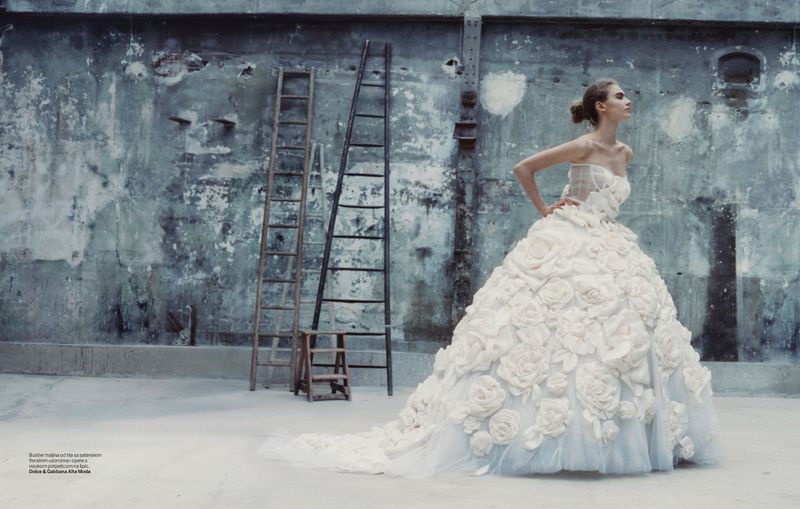 Woman in a white strapless floral gown, posing indoors on a grungy set with ladders. Fashion editorial photography, high-fashion concept.