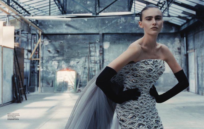 Fashion shot in industrial space featuring a woman in beaded dress with black gloves and grey train, standing in semi-profile view.