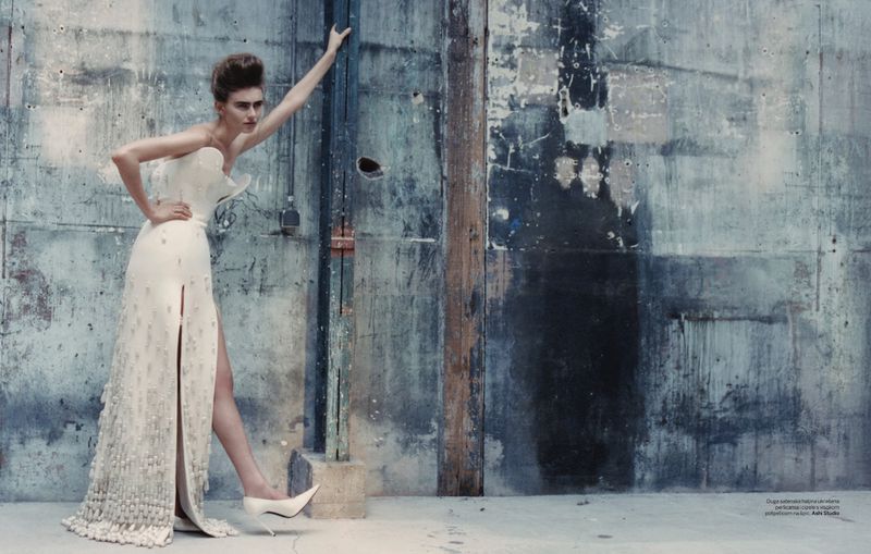Fashion editorial shot of a female model in a white couture gown posing against a distressed blue-gray textured wall in a studio.