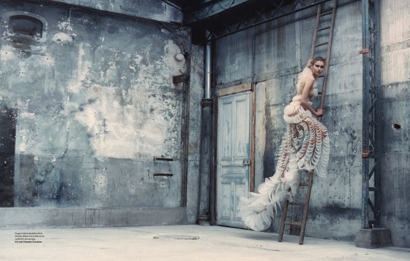 Woman in sculptural gown climbs weathered ladder in industrial interior, blending fashion with distressed architectural elements in a dramatic scene.