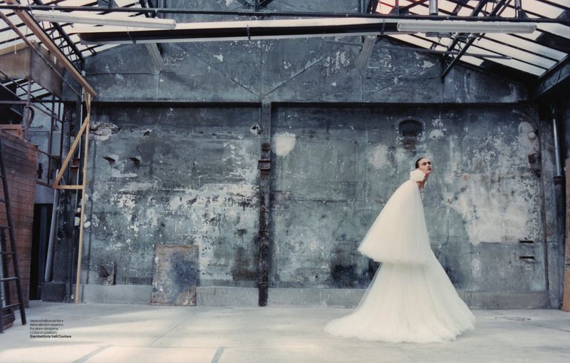 Woman in a white gown stands in a derelict warehouse with a grey wall, overhead lighting, and ladder on the left in full shot.