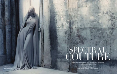 Model draped in a floor-length grey hooded gown leaning against a distressed industrial concrete wall