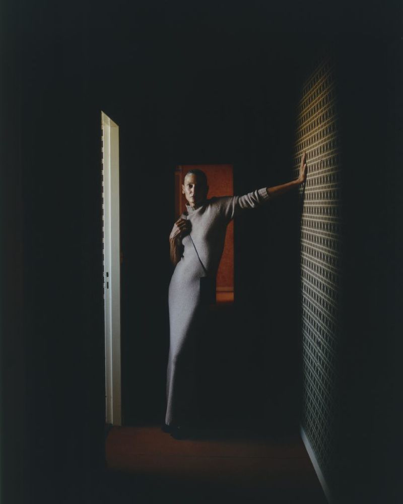 A woman in a gray dress poses in a dimly lit corridor with patterned wallpaper, partially open doors, and a red-toned end wall.