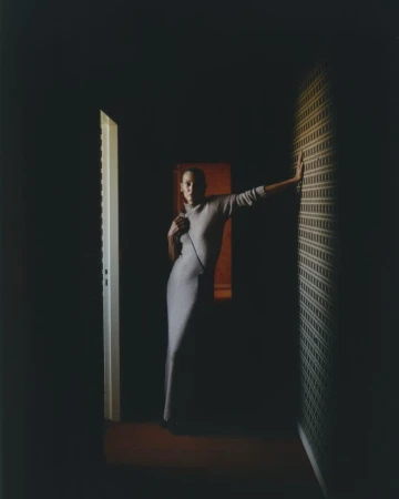 A woman in a gray dress poses in a dimly lit corridor with patterned wallpaper, partially open doors, and a red-toned end wall.
