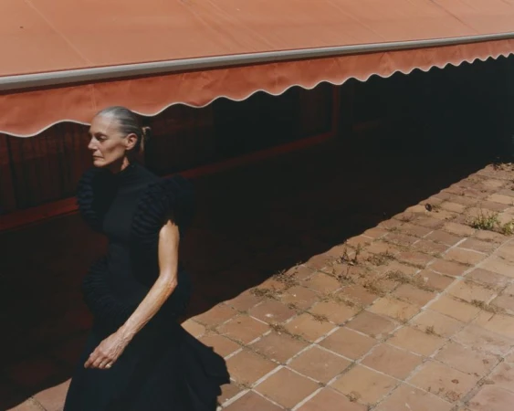Elegant older woman in black dress on a brick patio under an orange awning, captured in an eye-level shot, showcasing natural light.