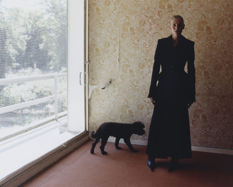 Elegant woman in a black coat poses with a dog indoors against patterned wallpaper, near a window with natural light.