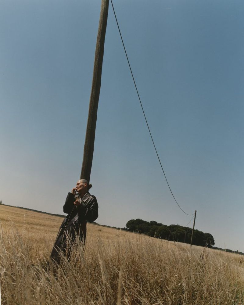 Photo of an adult standing near a telephone pole in a field of tall grass on a sunny day, wearing a dark coat.