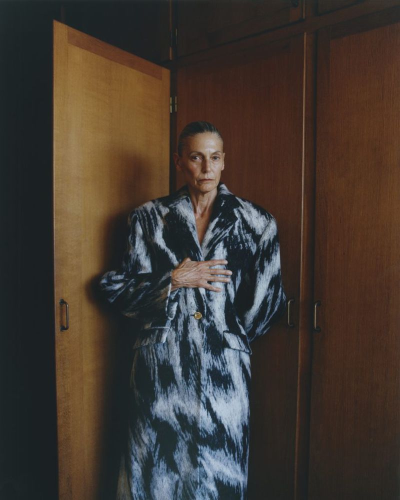 Older woman in a black-and-white coat poses indoors in front of a wooden wardrobe, looking directly at the camera with a neutral expression.