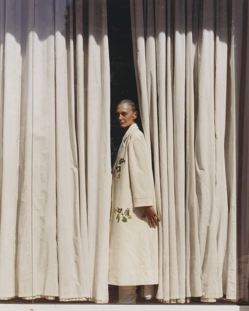 An older woman with short gray hair stands between white curtains in an eye-level shot, wearing a cream-colored coat with floral details.