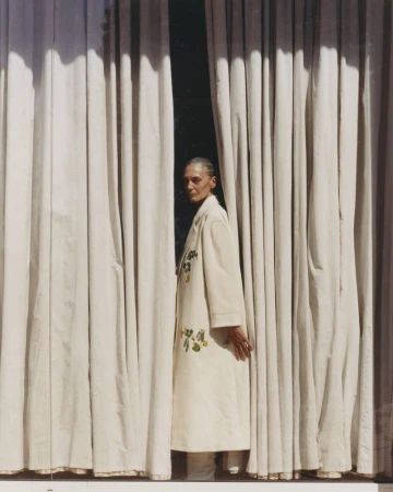 An older woman with short gray hair stands between white curtains in an eye-level shot, wearing a cream-colored coat with floral details.