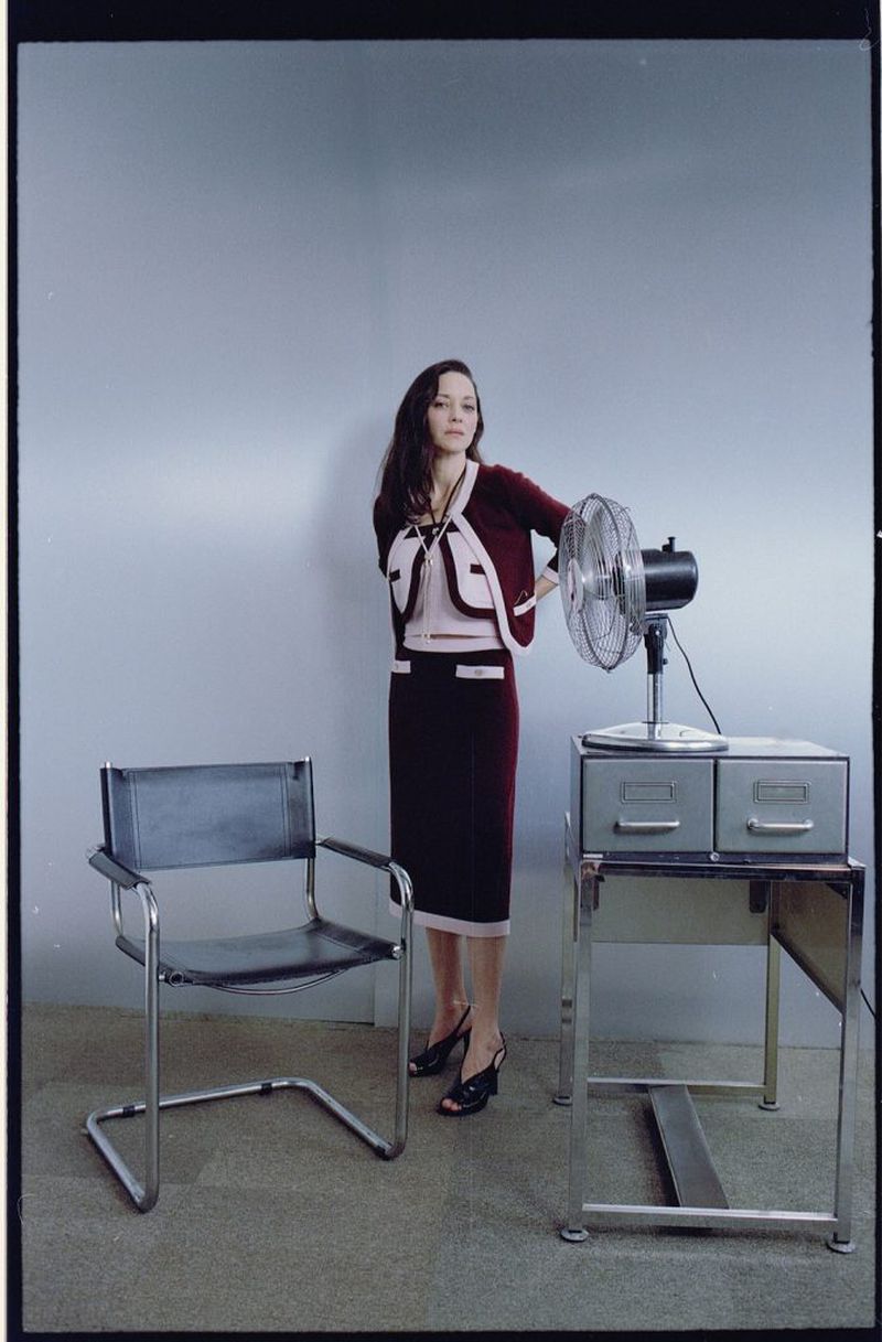 Brunette woman in a matching skirt and cardigan stands in front of a fan next to an office desk in a brightly lit interior.