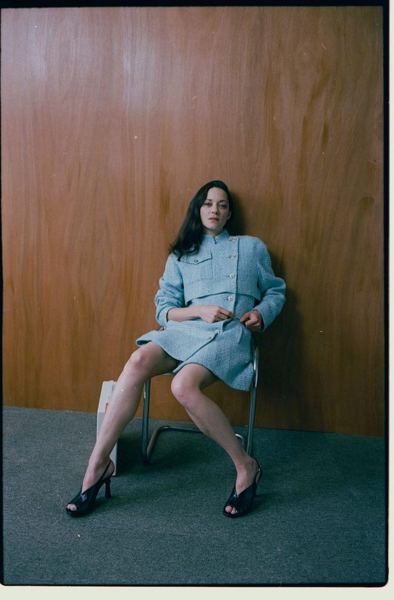 Adult female in an aqua tweed jacket and skirt sits on a metal chair against a wood panel wall, wearing black heels.