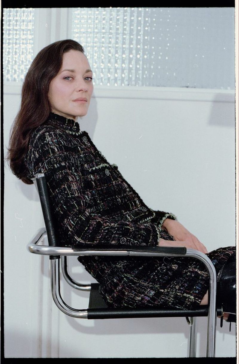 A woman with long brown hair is sitting in a metal chair in a stylish patterned jacket, posing in an indoor environment with a window.