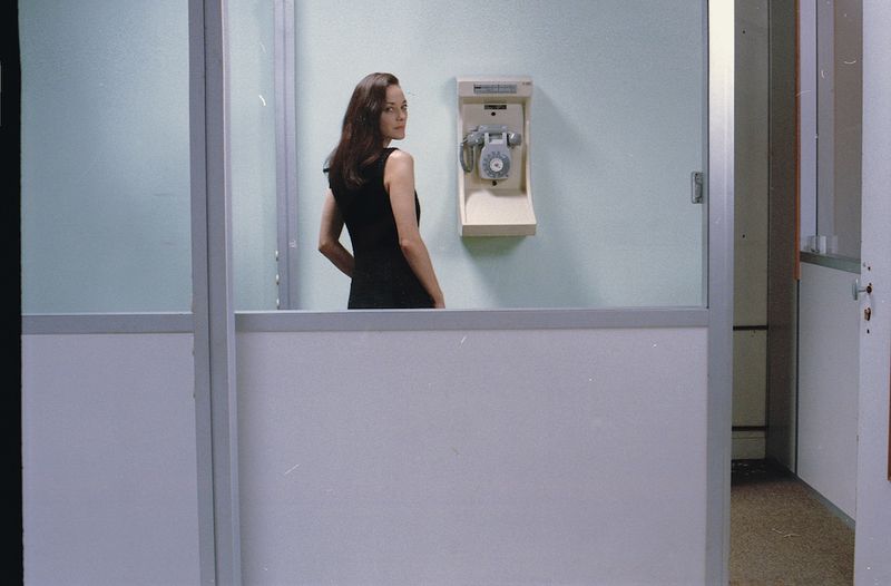 Brunette woman in a black dress stands inside an office space with glass partitions and an old-fashioned telephone mounted on the wall.