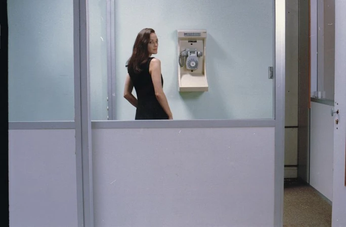 Brunette woman in a black dress stands inside an office space with glass partitions and an old-fashioned telephone mounted on the wall.