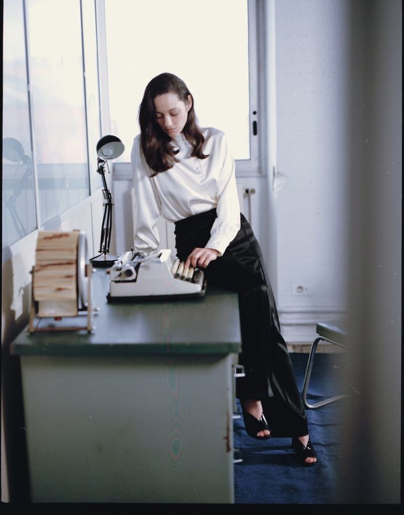 A woman in a white blouse types on a vintage typewriter while sitting on a desk in a light-filled interior, casting a focused gaze.