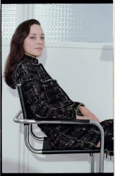 Marion Cotillard seated sideways in a chrome tubular armchair wearing a textured black tweed skirt suit against a white wall.