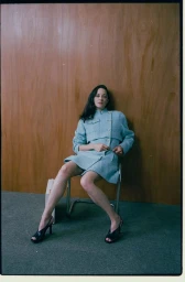 Marion Cotillard seated on a metal chair in a light blue tweed skirt suit against a wood panel backdrop.