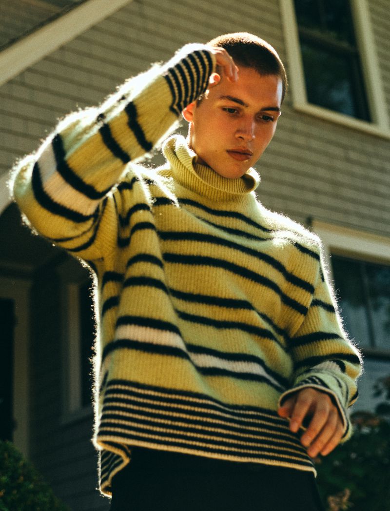 Young adult in a striped sweater stands outdoors in front of a house, gazing downward, fashion portrait, sunny day.