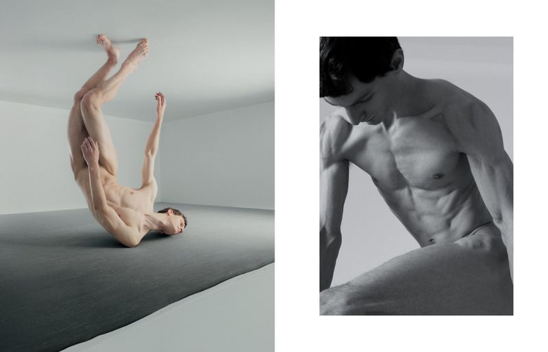 Athletic man in a studio with pale skin, dark hair, and visible muscle tone poses upside down against a grey floor and white wall.