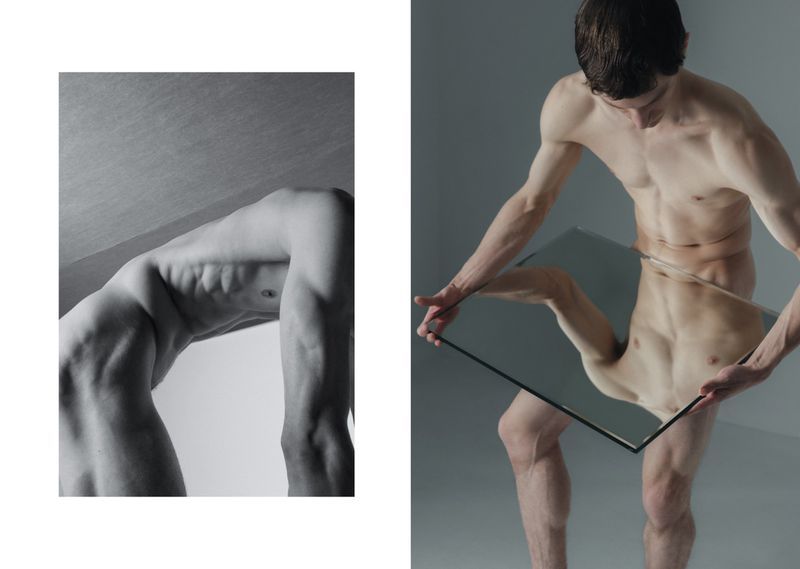 Two nude men in a studio. One man is bent over backwards and the other man is holding up a mirror, with reflected body parts.