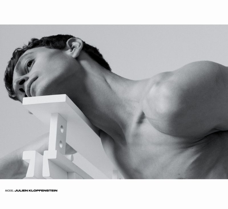 Black and white portrait of a shirtless man with dark hair leaning against a white, geometric structure in a light grey background.