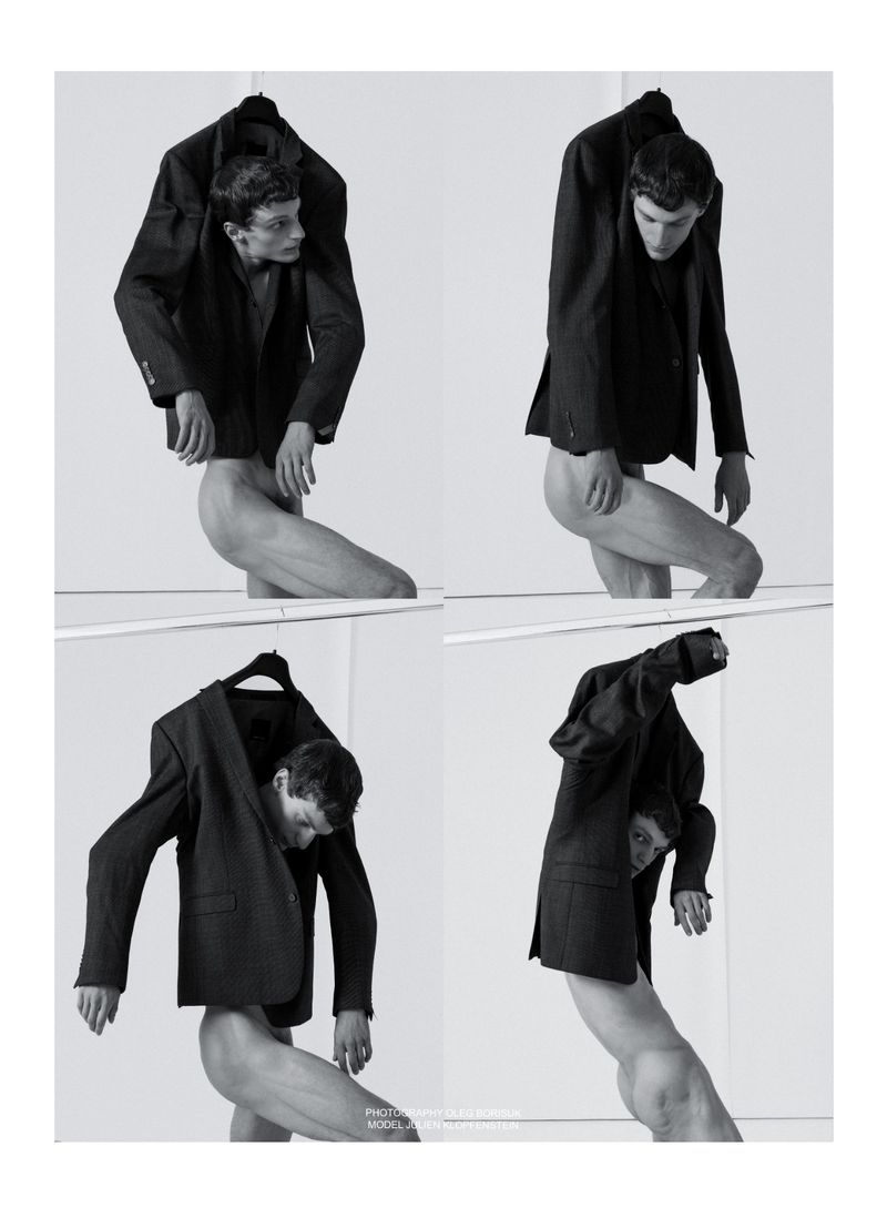 Conceptual black-and-white fashion shot featuring a nude male model wearing suit jackets, posed against a stark white backdrop in a photo studio.
