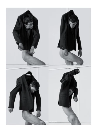 Conceptual black-and-white fashion shot featuring a nude male model wearing suit jackets, posed against a stark white backdrop in a photo studio.