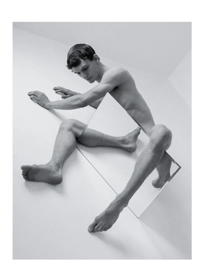 Monochrome studio portrait of a young man posed with a rectangular mirror intersecting his body on white seamless background.