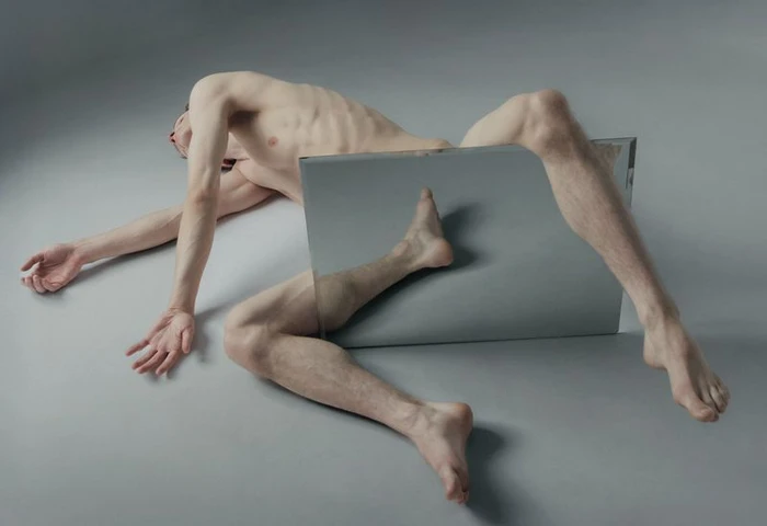 A male figure lies contorted on a grey studio floor beside a rectangular mirror reflecting his leg.
