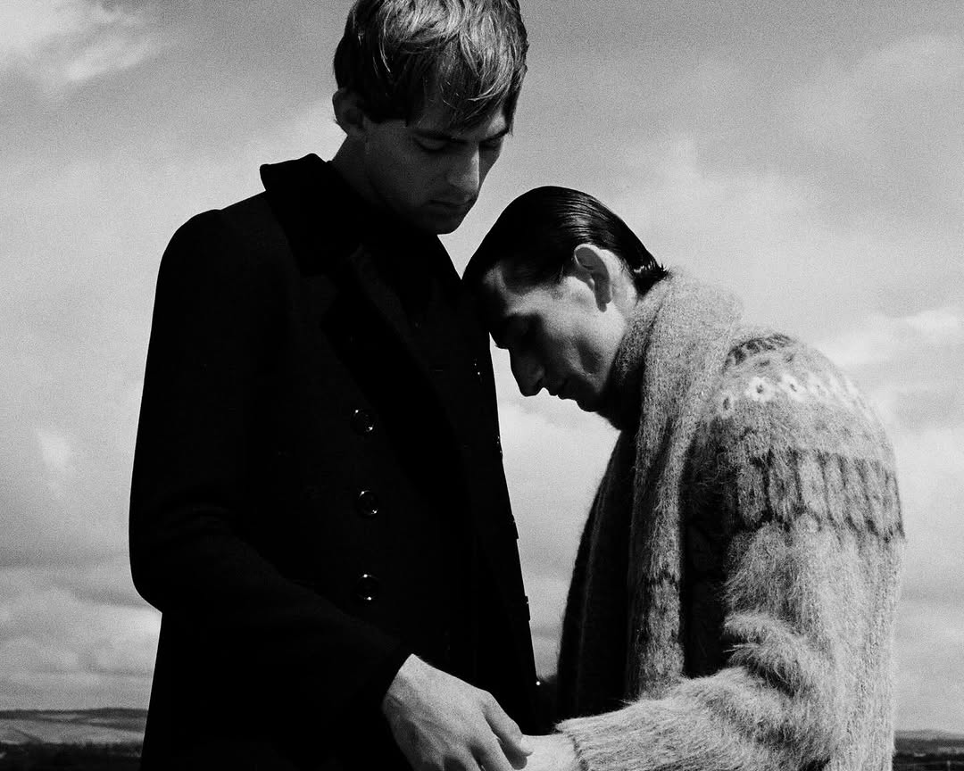 Monochrome portrait of two men in winter coats outdoors under an overcast sky, with one resting his head against the other.