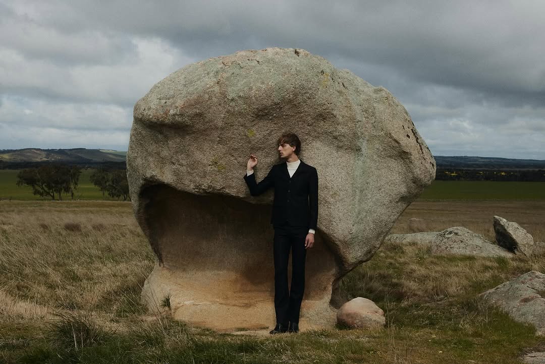 An adult man in a dark suit poses beside a large boulder in a grassy field under a cloudy sky in an outdoor editorial fashion shoot.