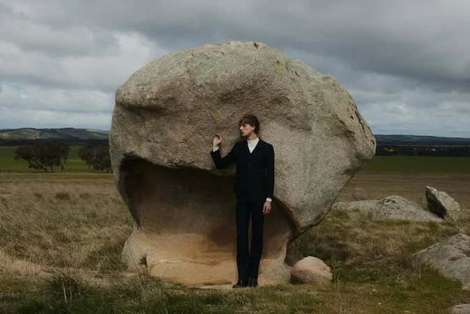 An adult man in a dark suit poses beside a large boulder in a grassy field under a cloudy sky in an outdoor editorial fashion shoot.