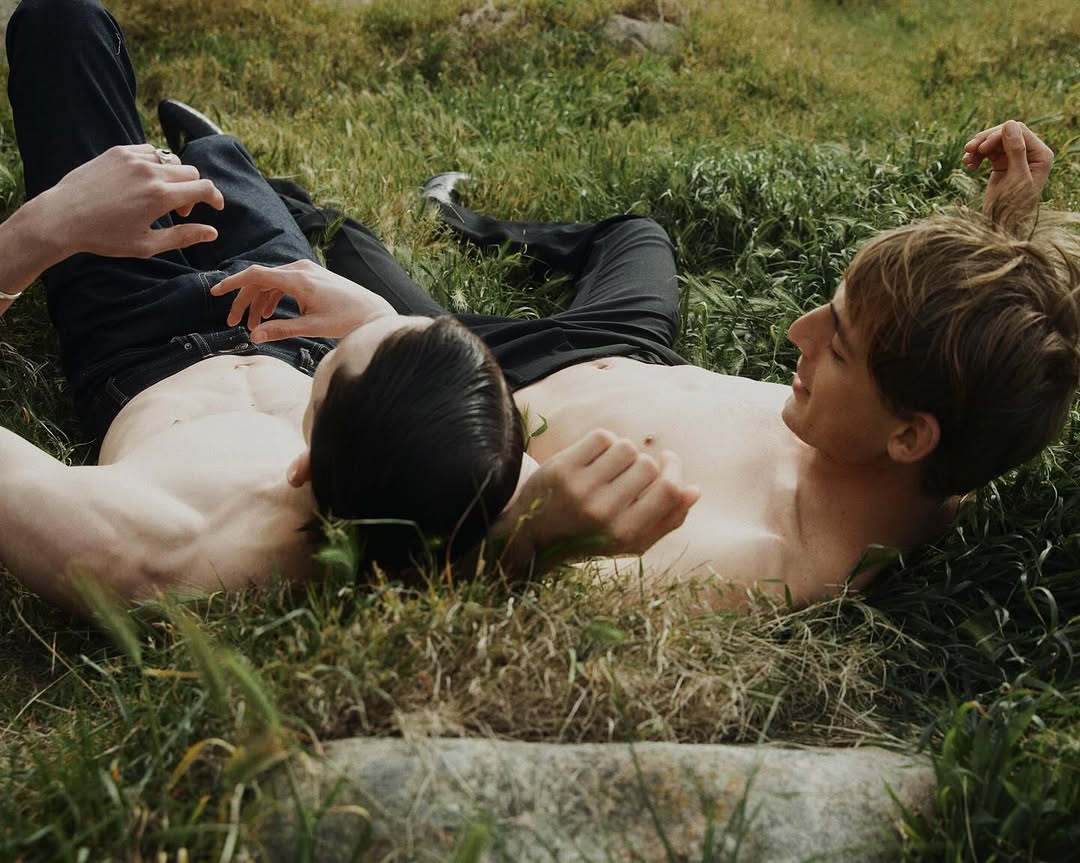 Two shirtless men in jeans lie on a grass lawn on a sunny day, their faces visible and their bodies partially exposed.