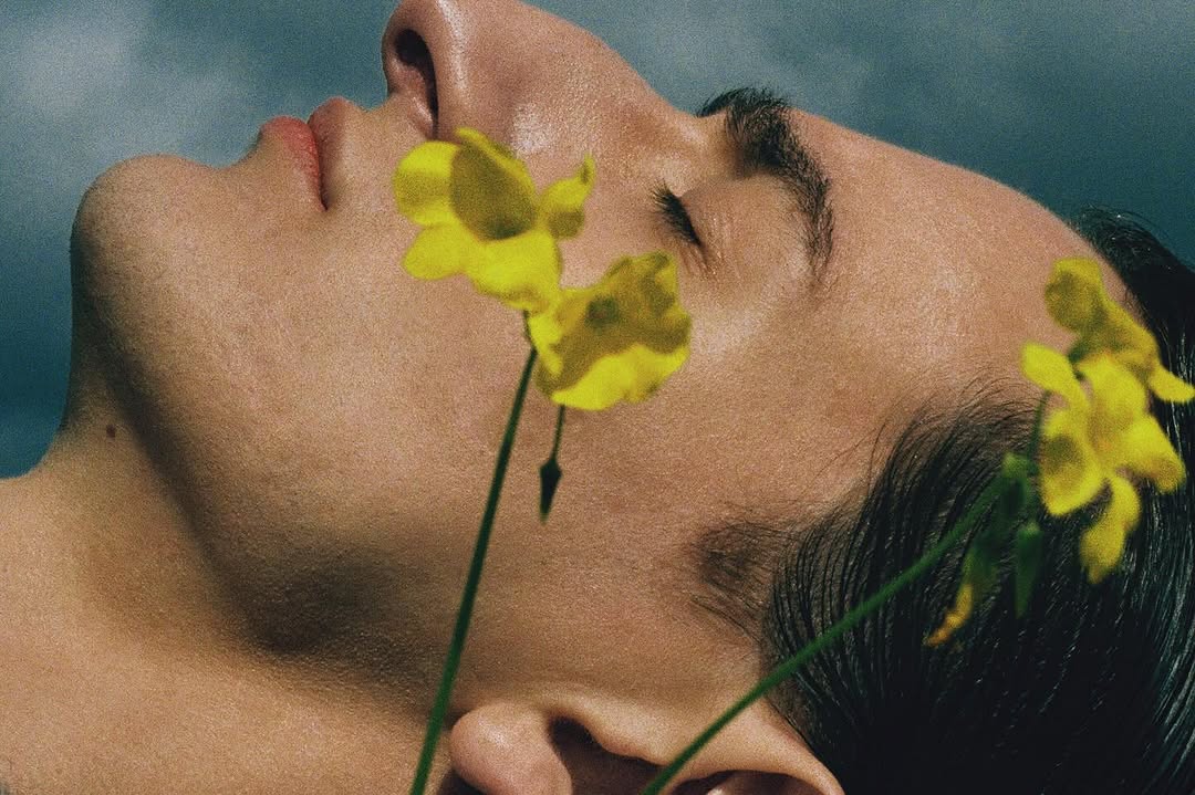Close-up portrait of a young adult male with fair skin, dark hair, and yellow flowers accenting a dreamy, contemplative mood.