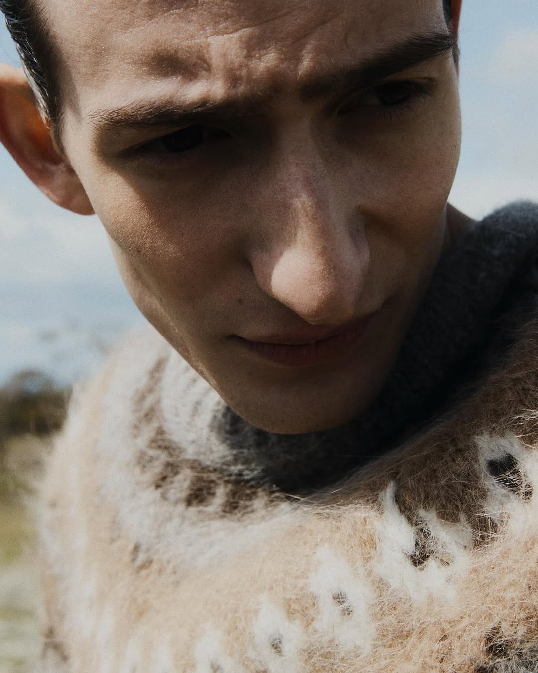 Close-up portrait of a young adult male with fair skin and short hair, wearing a patterned sweater, outdoors during the daytime.