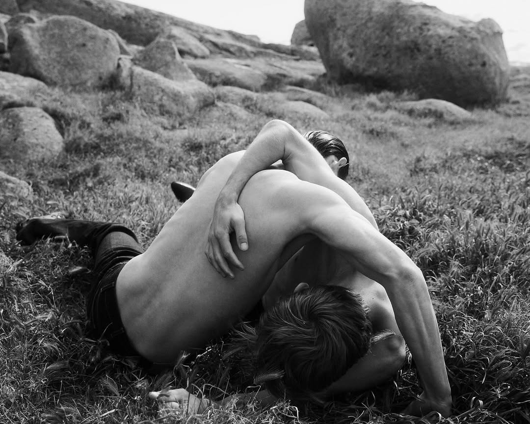 Black and white shot of two shirtless men wrestling on a grassy hillside surrounded by large boulders in an outdoor setting.
