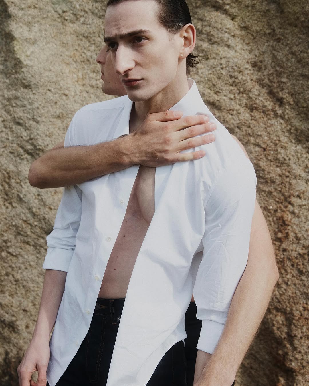 Two adult men pose closely outdoors with an unbuttoned white dress shirt against a textured rock formation backdrop.