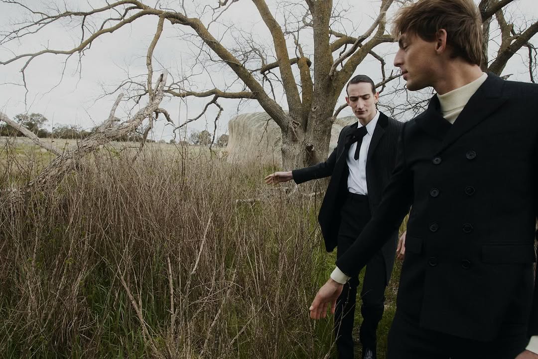 Two men in black suits pose outdoors in a field with a tree and hay bale. Fashion editorial shot. Muted color palette, low angle.
