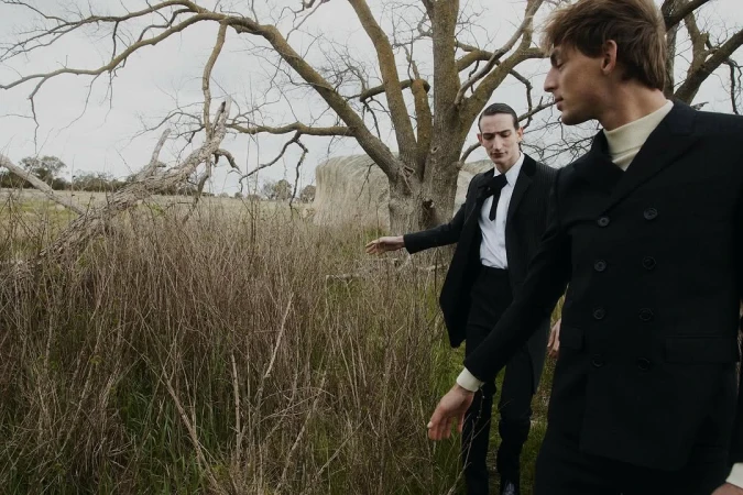 Two men in black suits pose outdoors in a field with a tree and hay bale. Fashion editorial shot. Muted color palette, low angle.