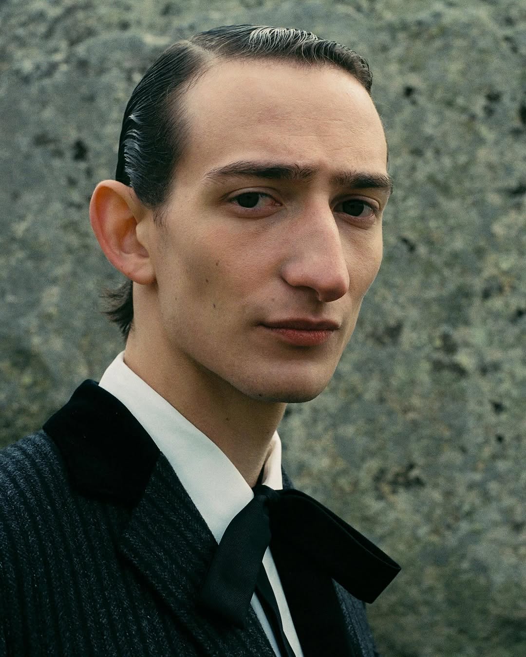 Close-up of a man with slicked-back hair, wearing a dark suit with a bow tie, posing against a textured outdoor background. 