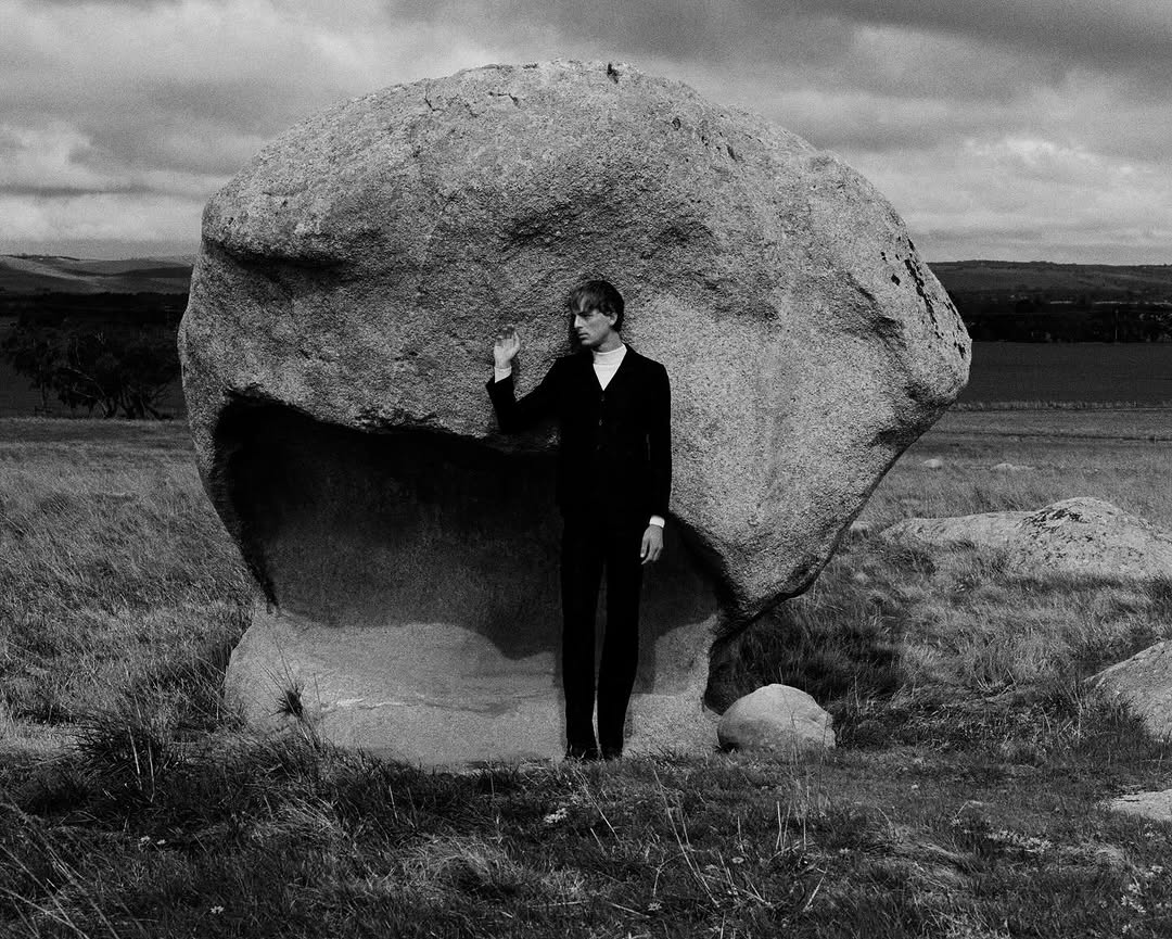 Monochrome portrait of a male figure standing next to a huge boulder in a grassy field under a cloudy sky, creating a surreal landscape.