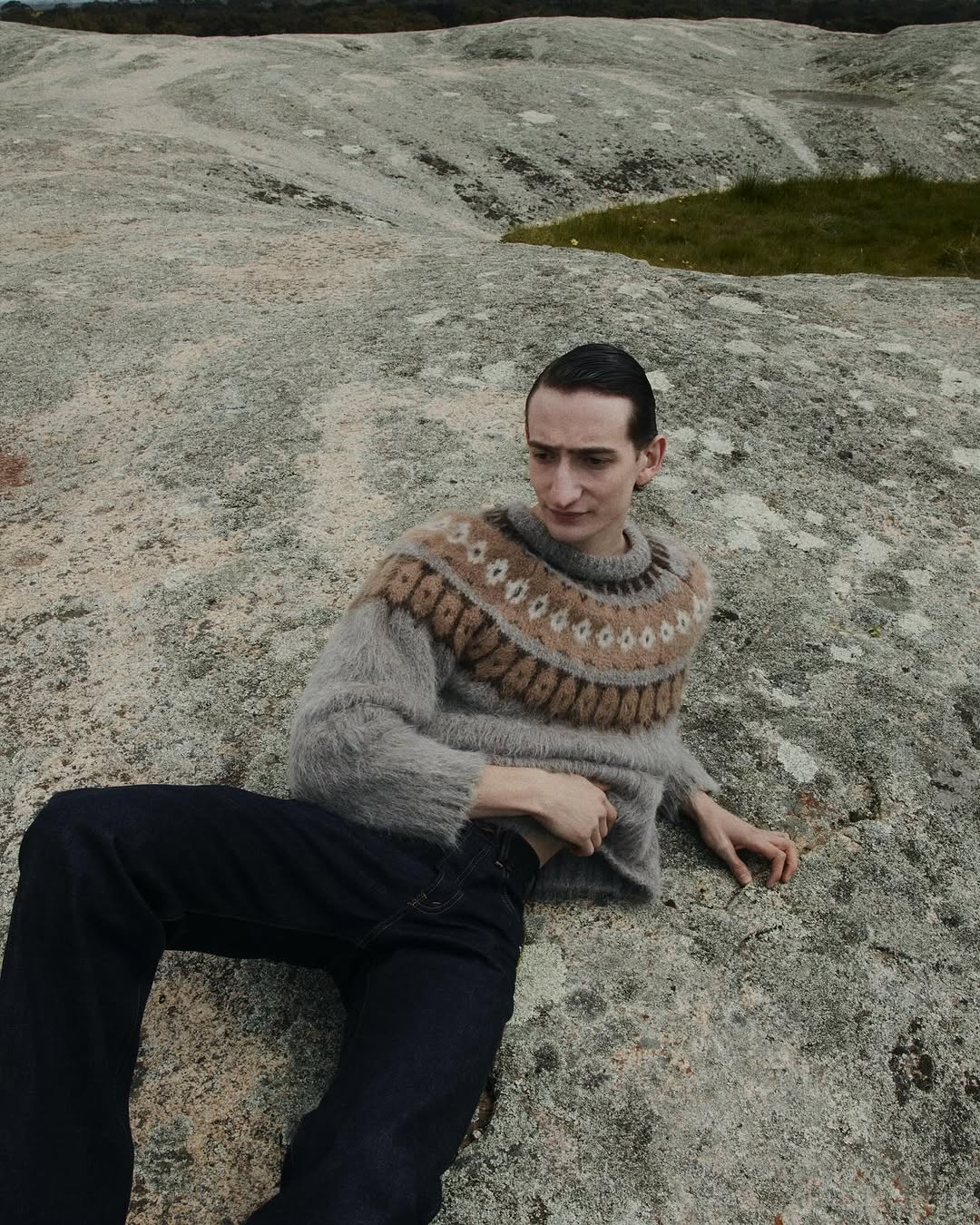 Young adult poses on a rock surface, wearing a grey and beige patterned sweater and dark jeans, set against a blurred natural backdrop.