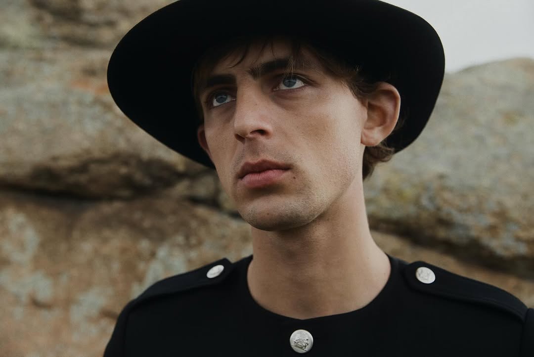 Portrait of a young adult male with fair skin wearing a black hat and black shirt posing in front of a rocky landscape in natural light.