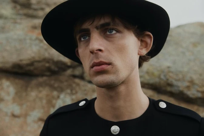 Portrait of a young adult male with fair skin wearing a black hat and black shirt posing in front of a rocky landscape in natural light.